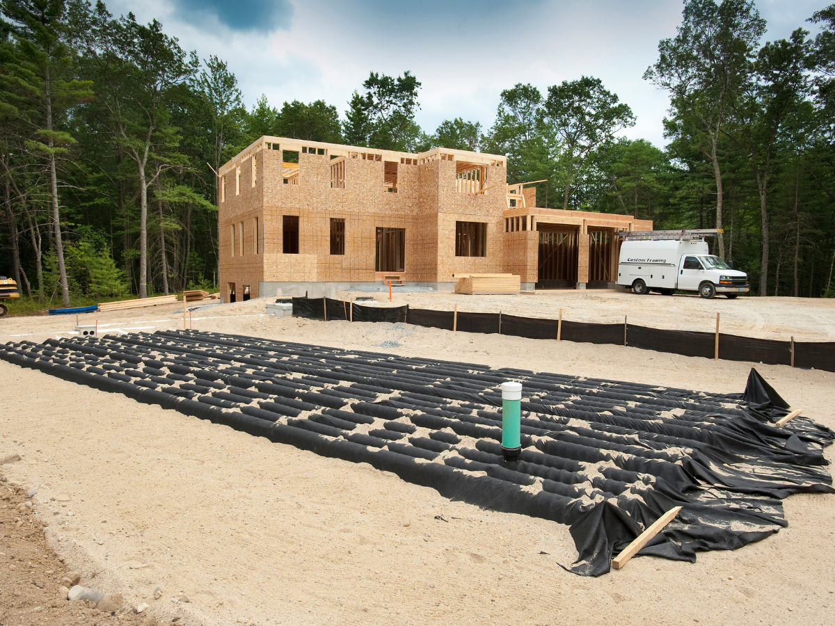 McCandless Plumbing & Excavation installs a new septic system for a house under construction in a sandy, tree-lined area.